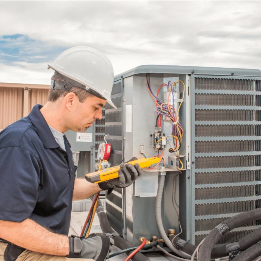 HVAC technician in a hard hat testing an outdoor air conditioner with a multimeter